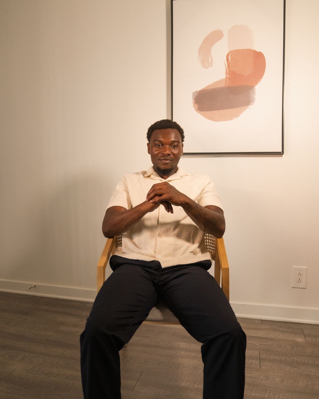 Portrait of a man in a cream textured shirt and navy trousers seated in a wooden cane chair, hands clasped, in front of a framed coral and peach abstract print