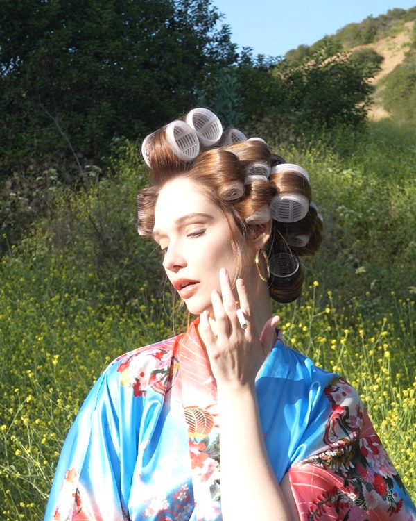 Woman in a blue and pink floral satin kimono with her hair in rollers, eyes closed and a hand at her cheek, in a hillside meadow of yellow wildflowers