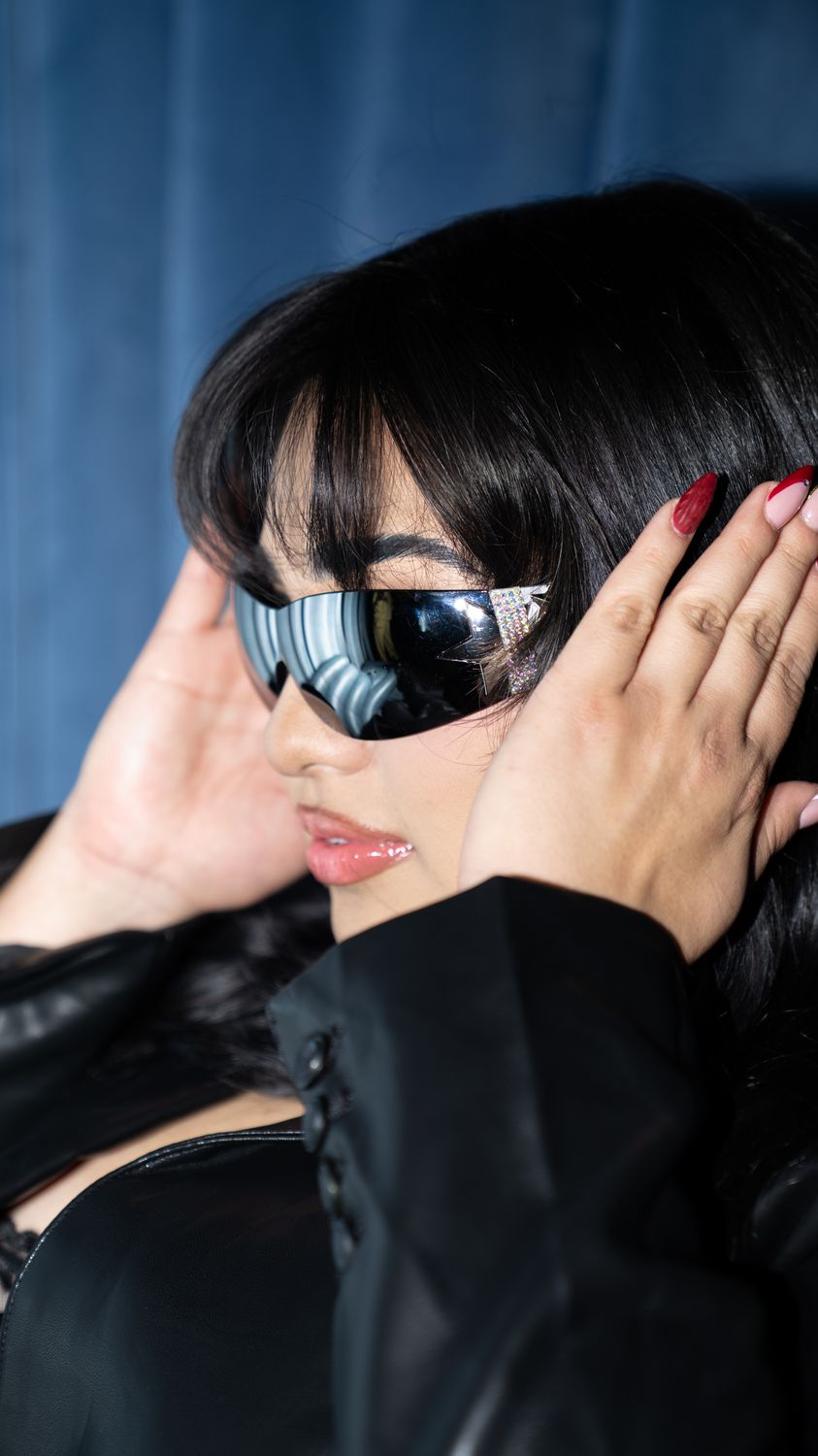 Close-up editorial portrait: mirrored wraparound sunglasses, hands framing the face against a blue backdrop