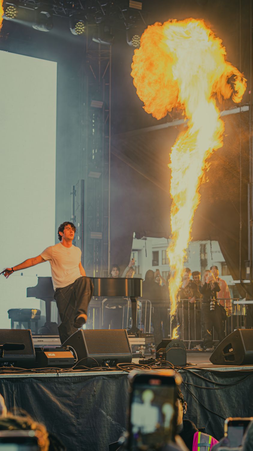 Performer mid-stride on a festival stage as a column of fire erupts beside a piano