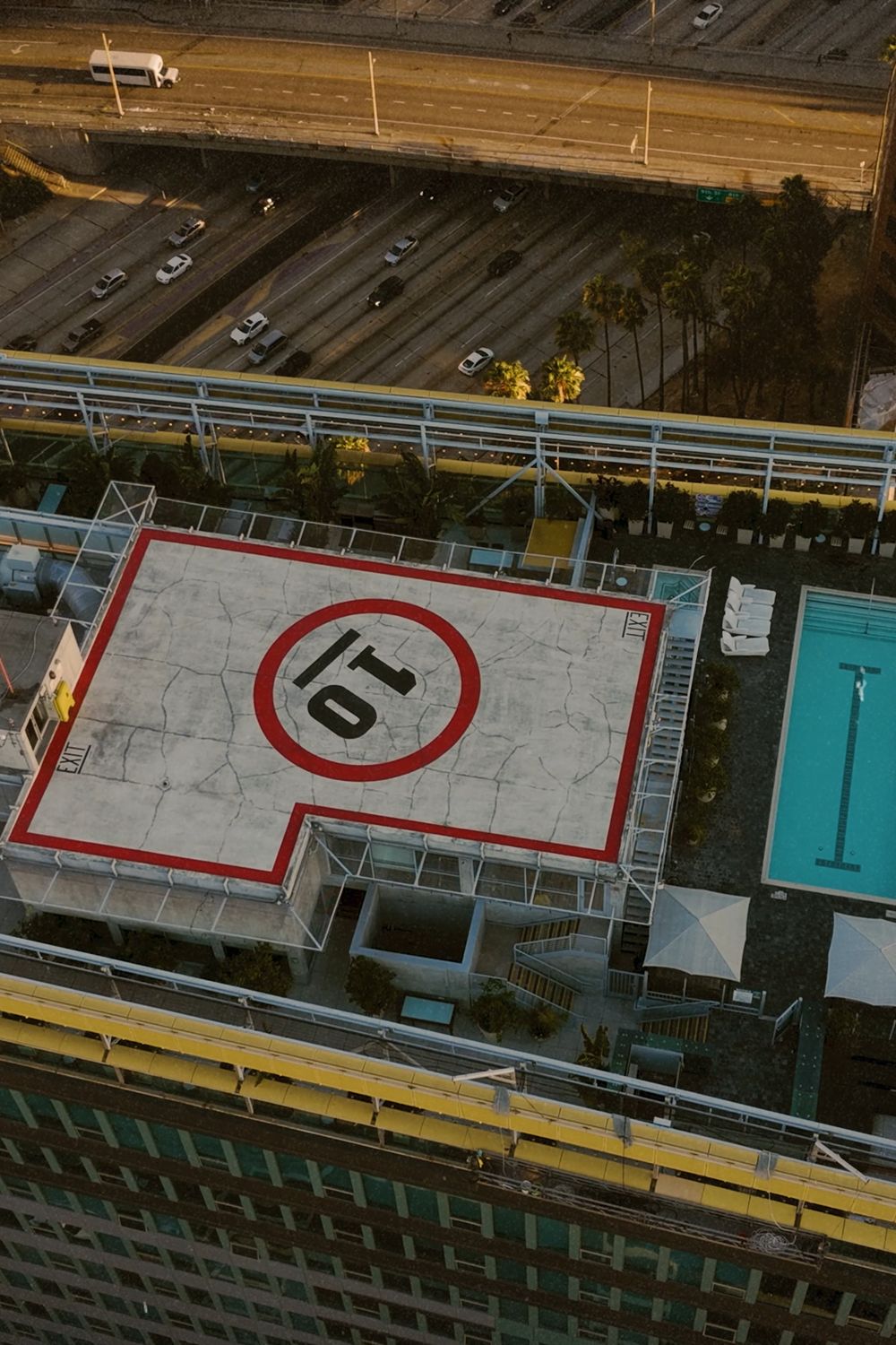 Top-down view of a red-ringed rooftop helipad beside a rooftop pool, a freeway above