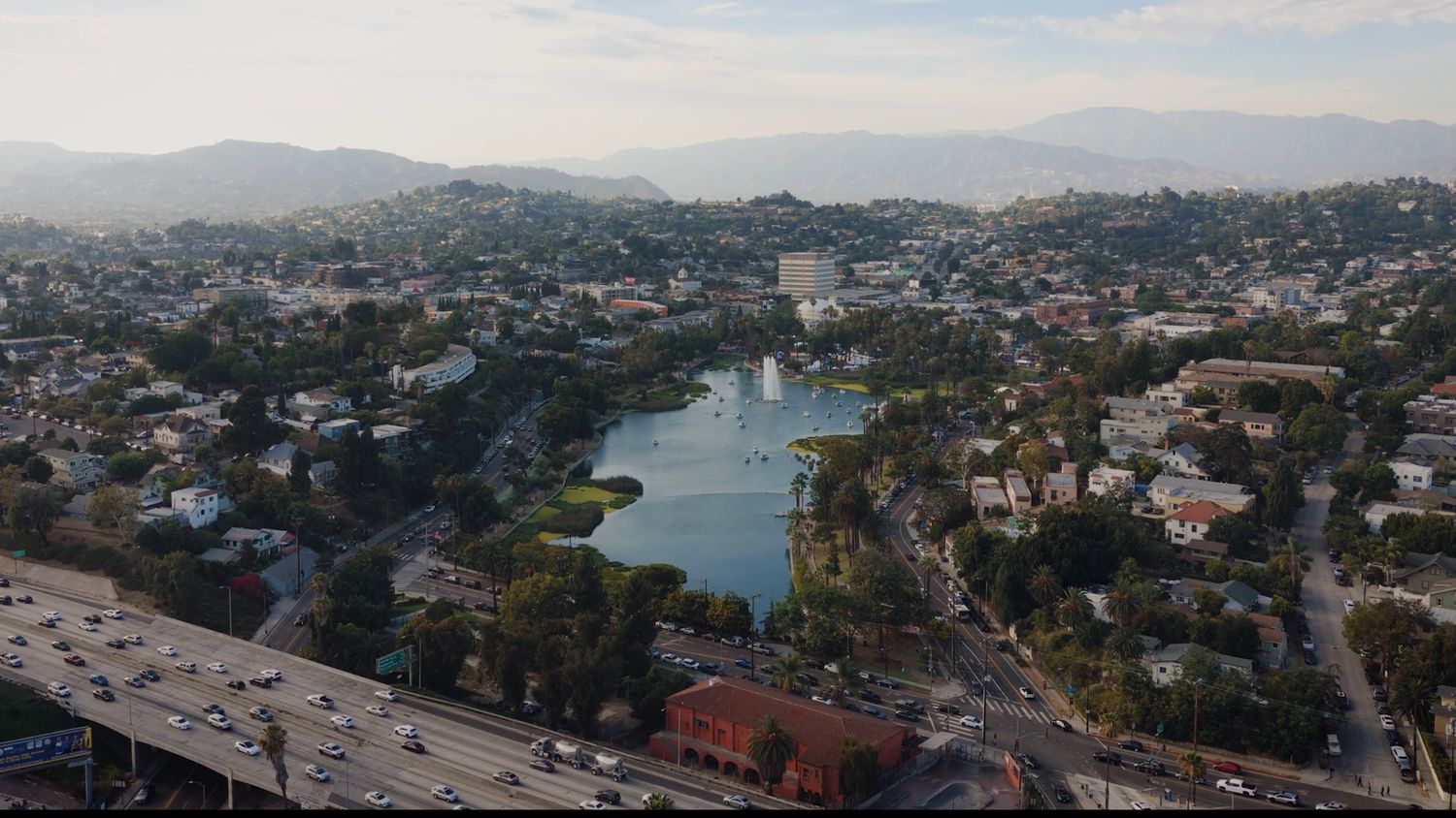 Aerial view over a lake with a fountain, hillside neighborhoods around it, a freeway in the foreground and mountains beyond