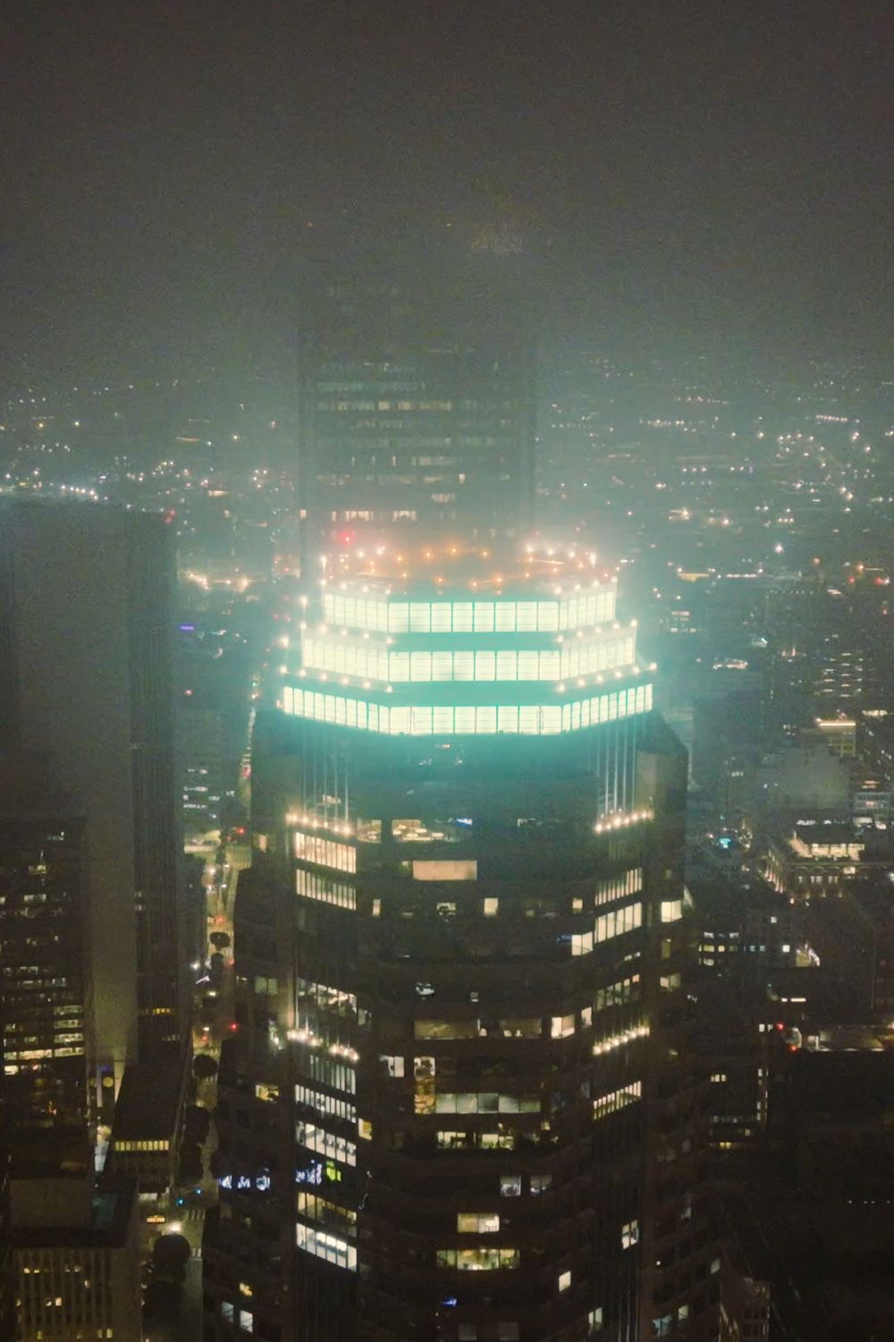 Aerial view at night of a crown-lit downtown tower rising through haze above the city grid