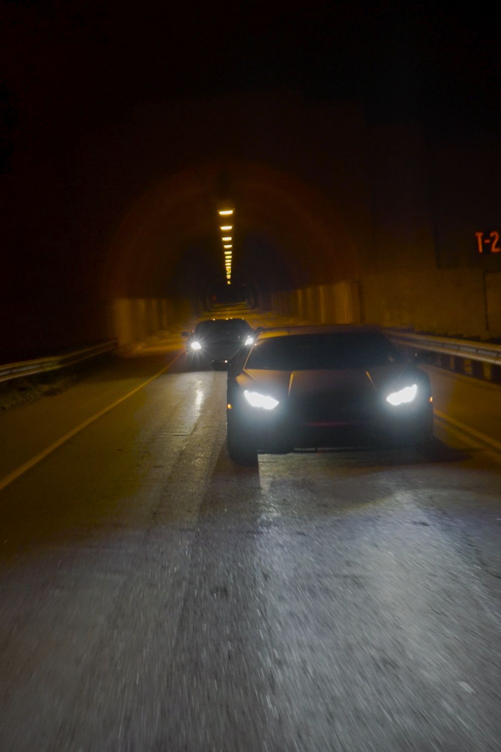 A sports car with its headlights on driving toward the camera through a tunnel lit by amber lamps