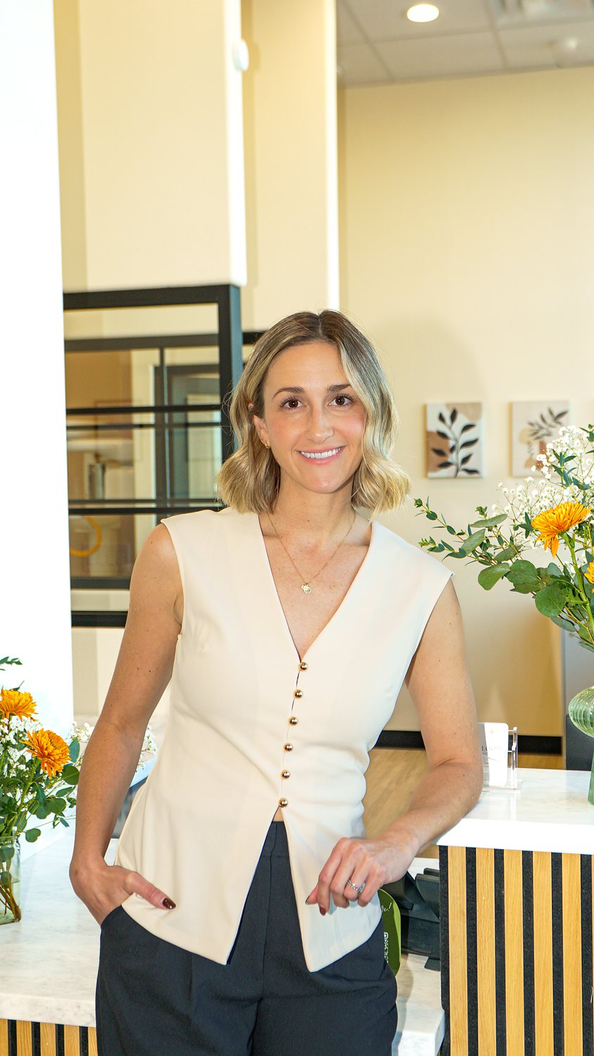 A smiling woman in a cream vest and dark trousers leans on a reception counter beside a bouquet of orange and white flowers in a bright dental practice