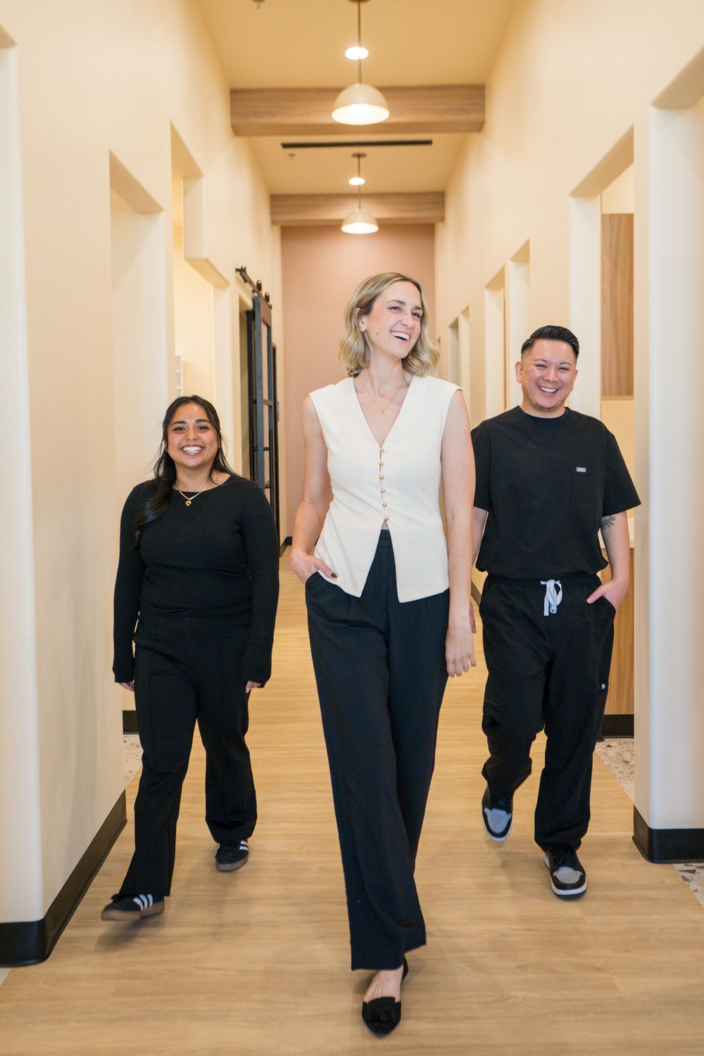 A dentist in a cream vest walks down a bright corridor between two team members in black scrubs, laughing