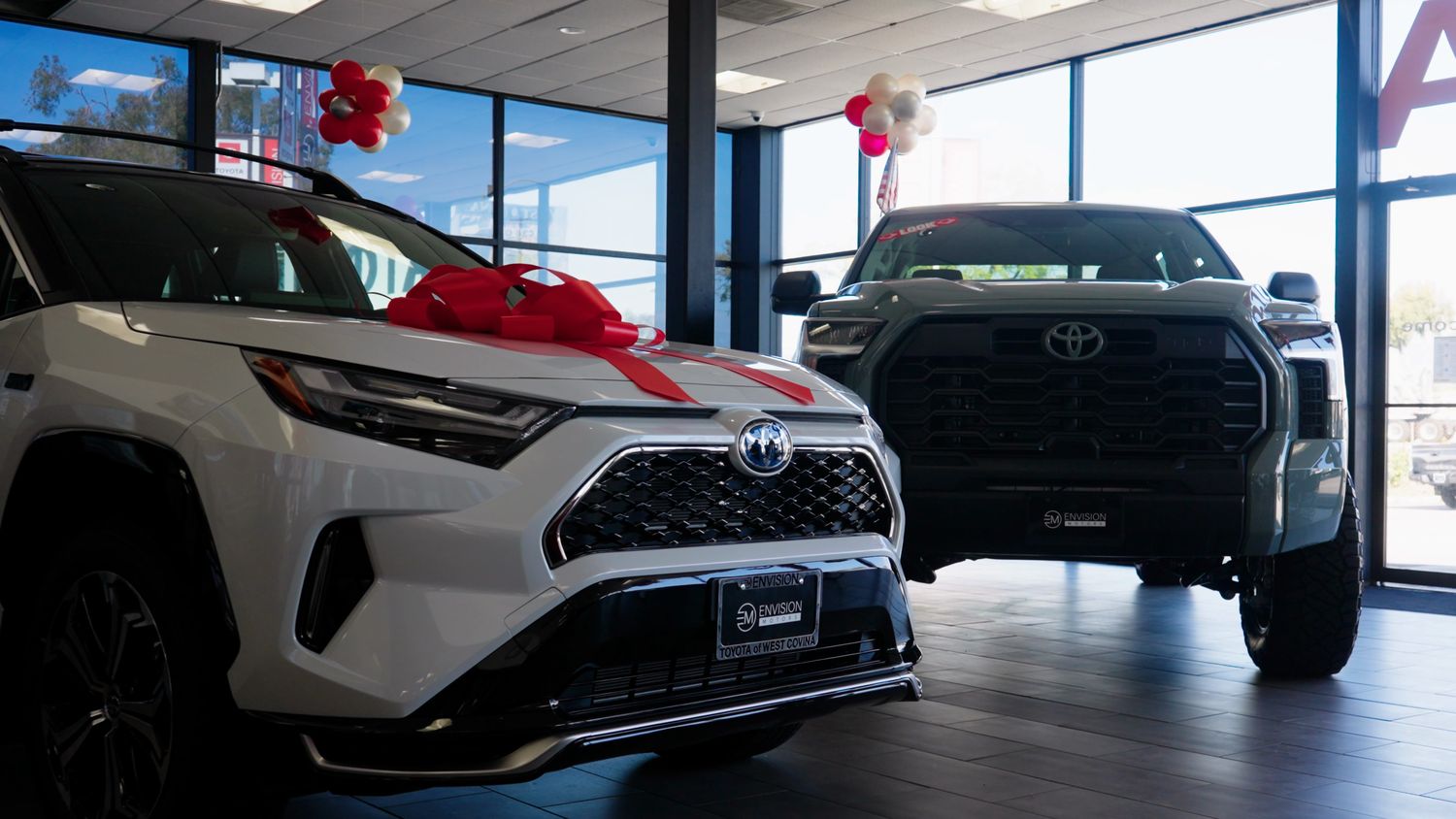 A white hybrid SUV wearing a red bow beside a gray pickup in a bright showroom with tall windows and balloons