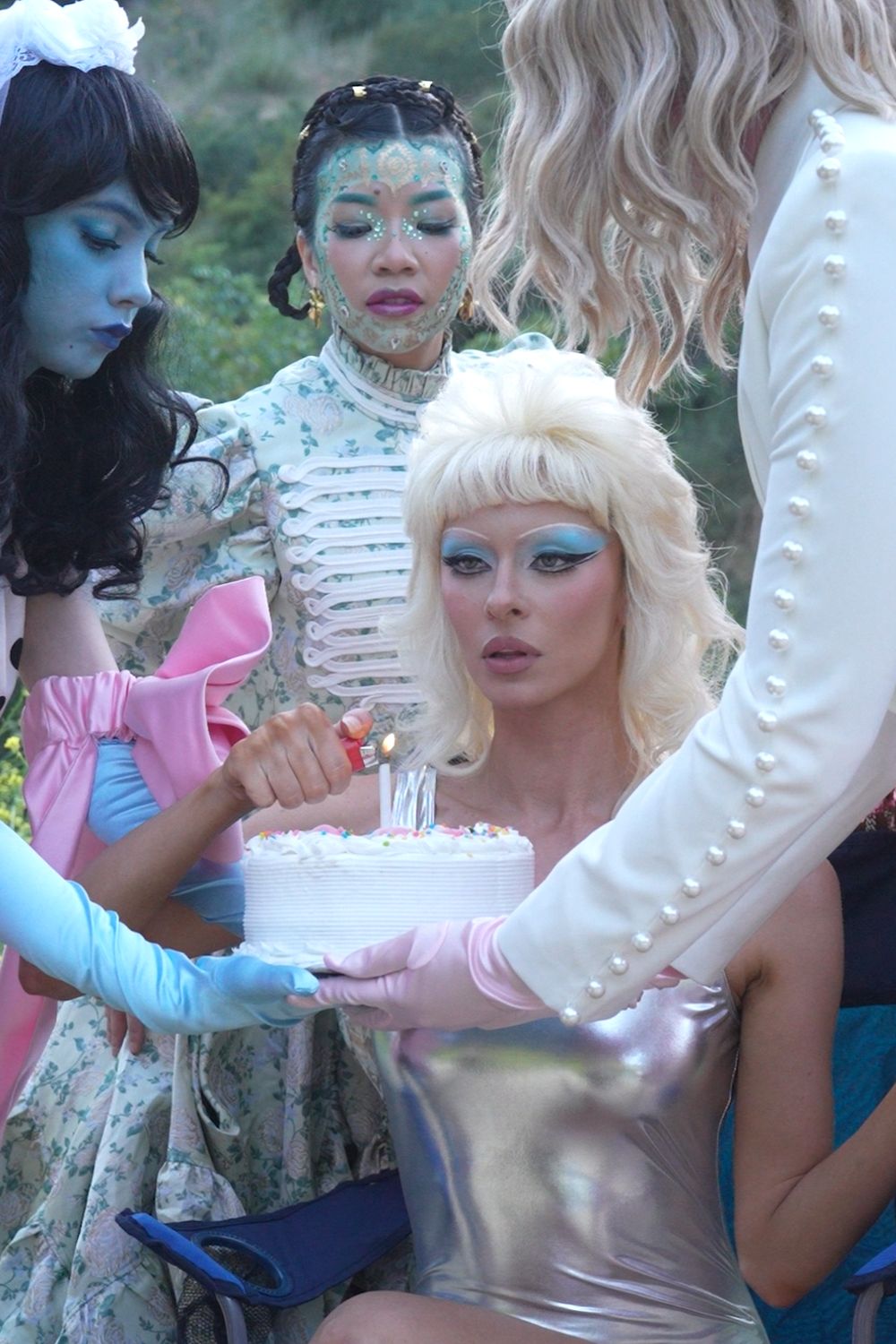 Fashion tableau on a hillside: models in pastel couture and face paint gather around a small white cake as one lights the candle