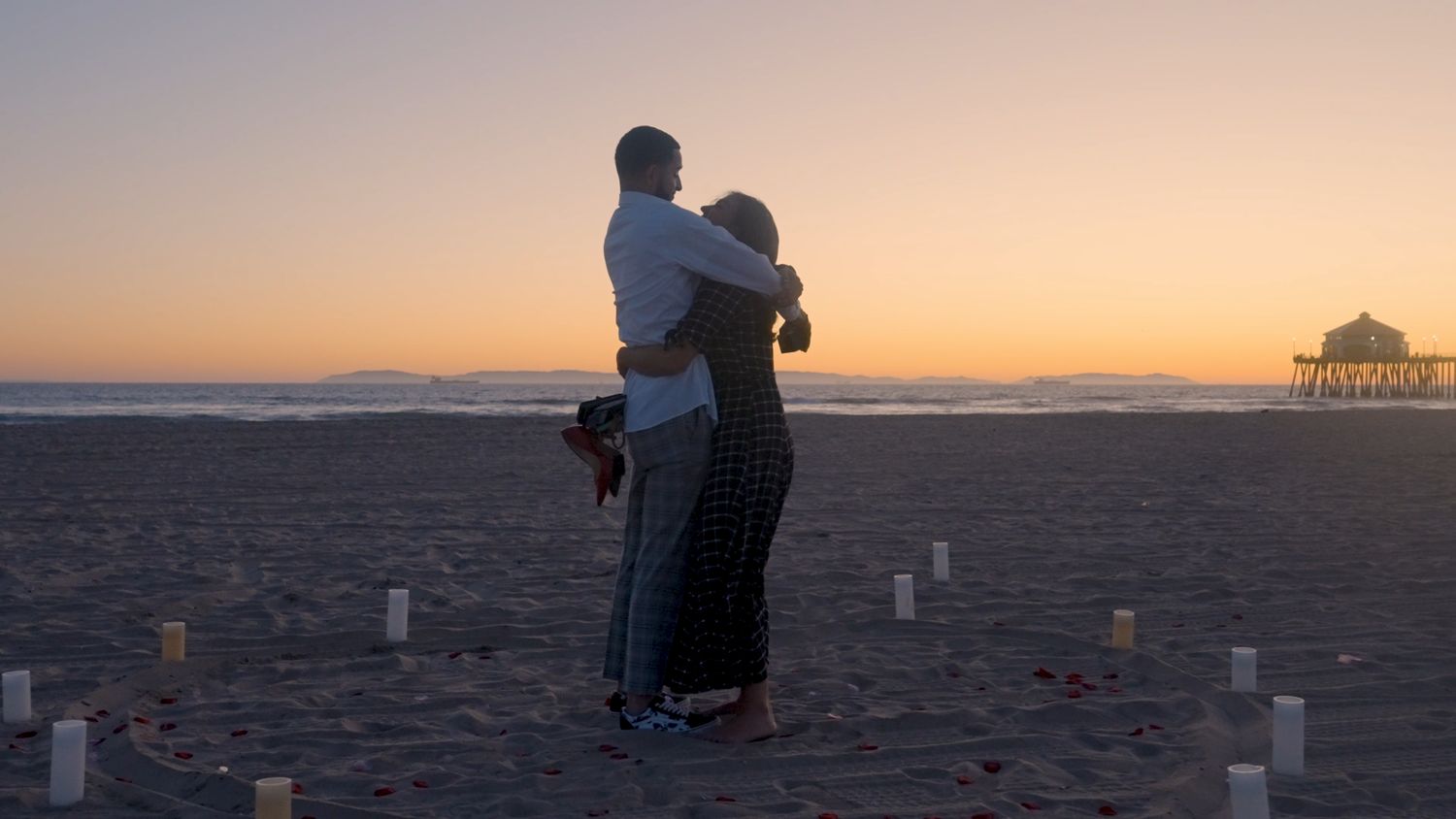 A couple embraces on a beach at dusk among candles and rose petals, a pier on the horizon