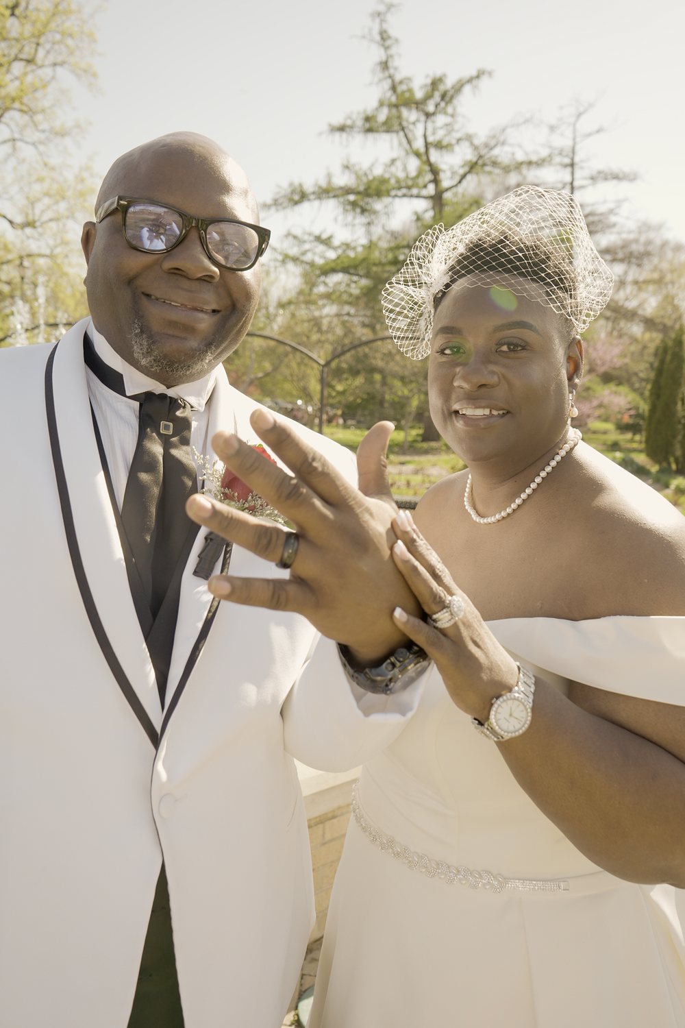A bride in a birdcage veil and a groom in a white tuxedo jacket smile and raise their hands to show their wedding rings in a sunlit garden