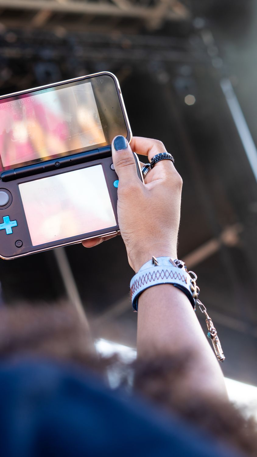 A hand holds a handheld game console toward a festival stage, its twin screens glowing with stage light