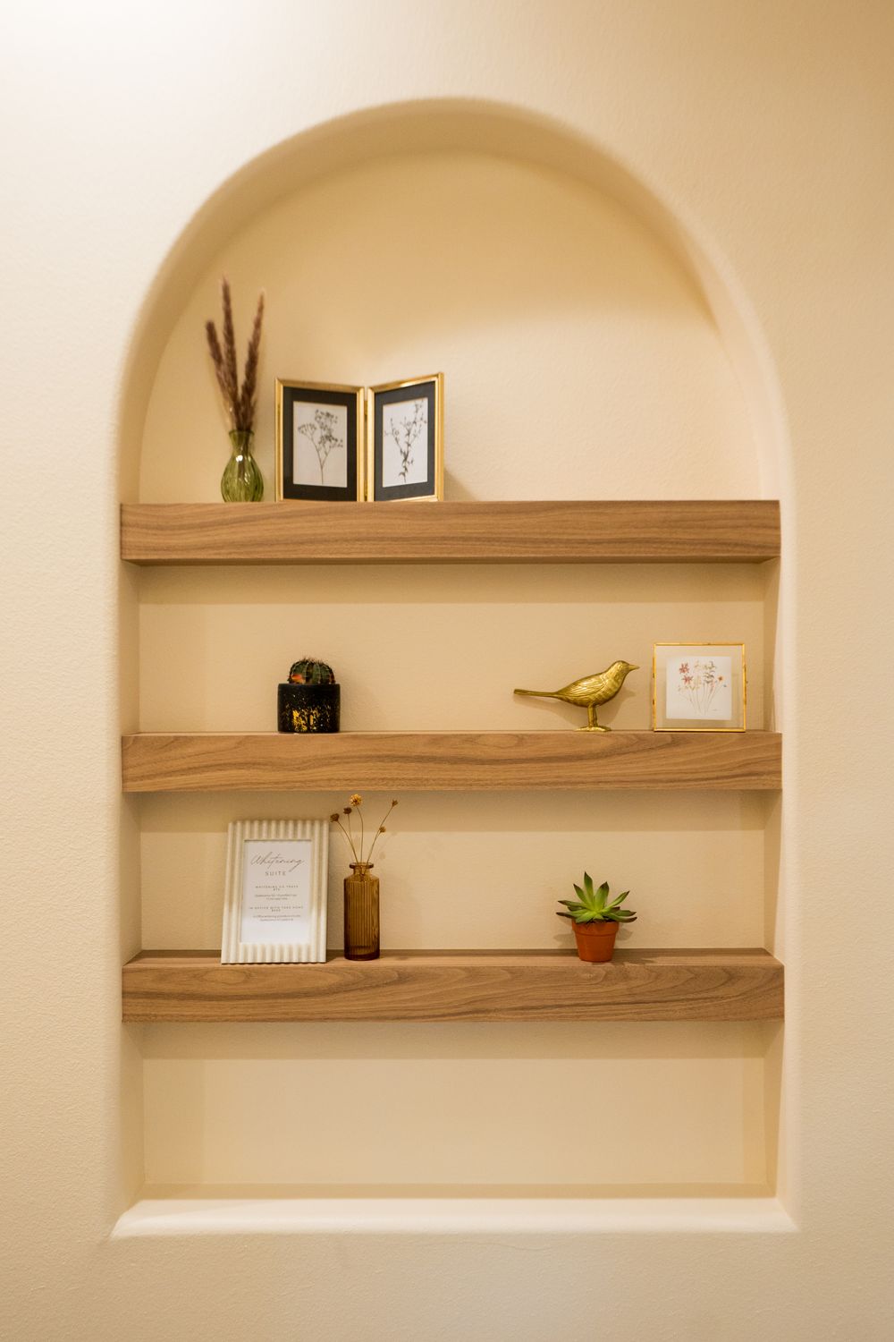 An arched plaster niche holding three wooden shelves with framed botanical prints, a brass bird and small plants