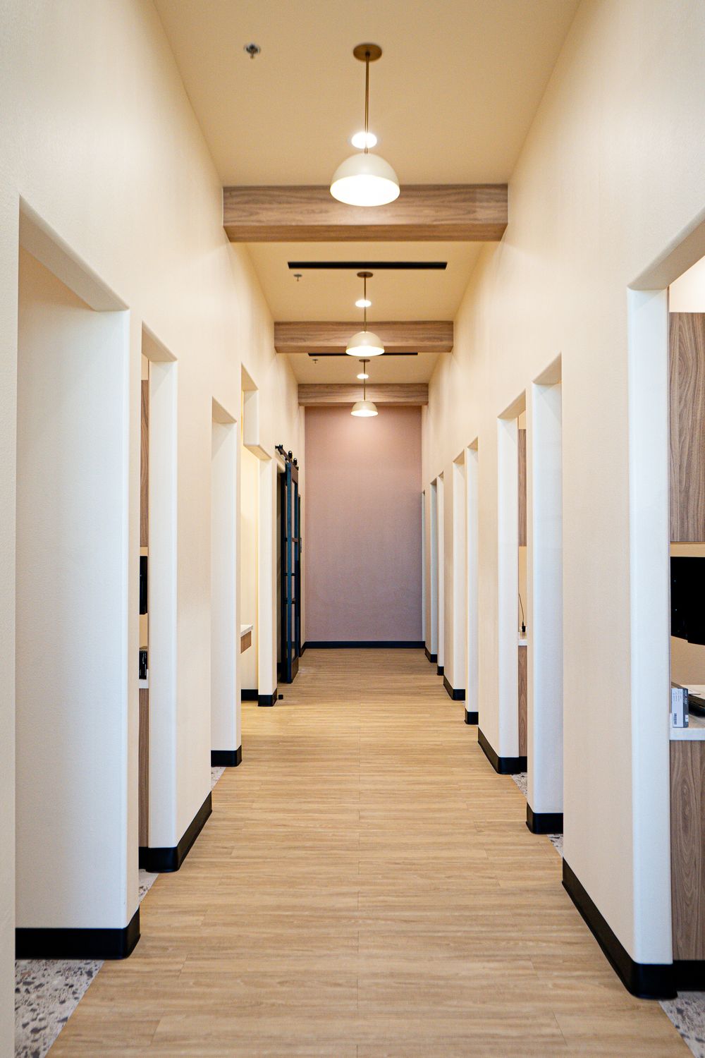 A long, sunlit clinic corridor with pendant lamps, warm wood beams and a blush-pink wall at the far end